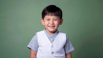 小男孩穿着白色背心和灰色短袖面带笑容站在绿色背景前的图片1