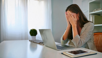 一位坐在桌前面对笔记本电脑用手掩面表现出哀伤情绪的女性