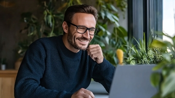 戴眼镜的男子在绿植环绕的室内使用笔记本电脑微笑