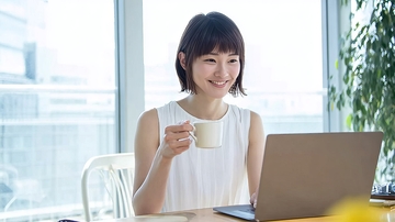 年轻女性在明亮办公室使用笔记本电脑并手持咖啡杯